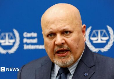 International Criminal Court Prosecutor Karim Khan speaks during an interview while sitting in front of a blue sign with blurry white writing and the symbol of the ICC - the scales of justice surrounded by a laurel wreath. 