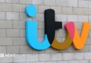 Mobile phone with an ITV 1 logo in black and yellow on the screen and blurred images of a streaming service in the background