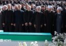 Iranian President Masoud Pezeshkian, Iran's Parliament speaker Mohammad Baqer Qalibaf and Chief Justice of Iran Gholam-hossein Mohseni Ejei with officials attend a prayer during a public farewell ceremony to pay their respects to late Supreme Leader Ayatollah Ali Khamenei. A large crowd can be seen in the background as everyone stands looking at the coffin
