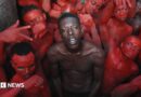 A topless Empereur stands in the centre of a group of young men, who are painted in red.