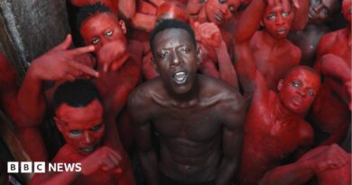 A topless Empereur stands in the centre of a group of young men, who are painted in red.
