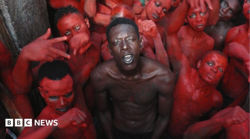 A topless Empereur stands in the centre of a group of young men, who are painted in red.
