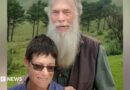 Rodney Saunders stands behind Rachel. Rodney has a long grey beard while Rachel has dark hair and is wearing sunglasses