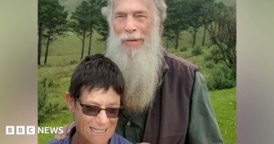 Rodney Saunders stands behind Rachel. Rodney has a long grey beard while Rachel has dark hair and is wearing sunglasses