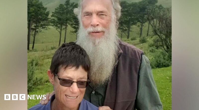 Rodney Saunders stands behind Rachel. Rodney has a long grey beard while Rachel has dark hair and is wearing sunglasses