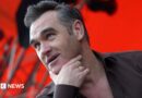 Head and shoulders of Morrissey on stage with his right hand thumb and index finger cupping his chin. He is wearing a gold signet ring, a black shirt and the background is lit red with scafolding