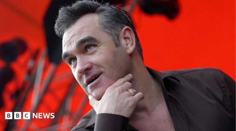 Head and shoulders of Morrissey on stage with his right hand thumb and index finger cupping his chin. He is wearing a gold signet ring, a black shirt and the background is lit red with scafolding
