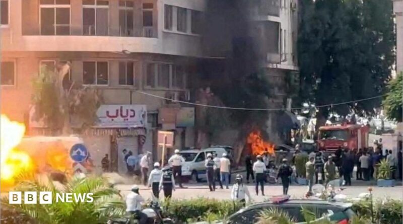 A wide shot of a street with lots of emergency workers. Flames and smoke rise from an alleged explosion site