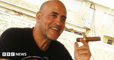 Daniel Siad, a man with a shaved head and stubble, wearing a black T-shirt, sits outside in what appears to be a sunny street, smiling with a large cigar raised in his hand.