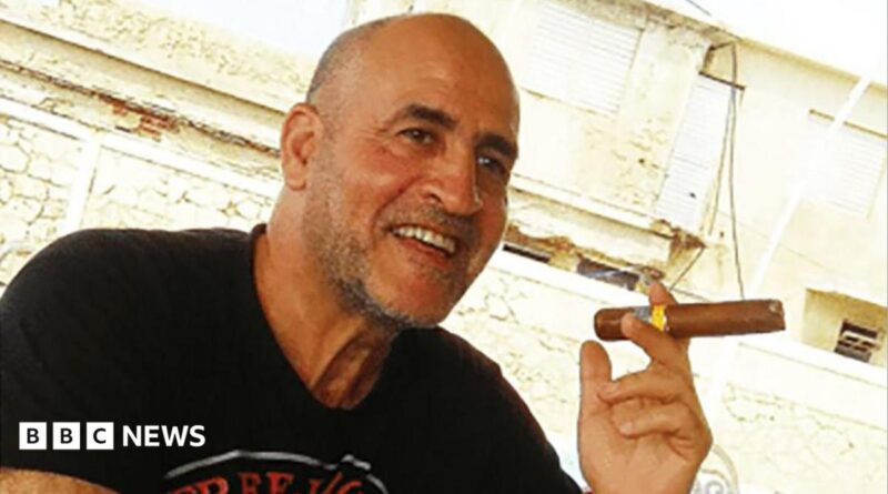 Daniel Siad, a man with a shaved head and stubble, wearing a black T-shirt, sits outside in what appears to be a sunny street, smiling with a large cigar raised in his hand.