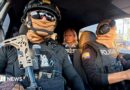 Two Ecuadoran police officers in dark sunglasses, helmets and balaclavas - which entirely obscure their faces - sit in a car in front of the BBC's Orla Guerin, wearing a flak jacket with the word "PRESS" on it. 