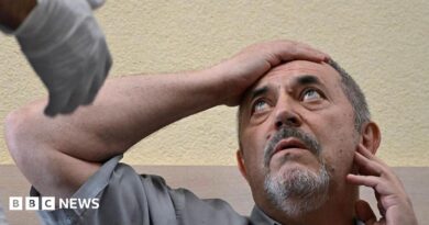 A man in a grey shirt holds his right arm to his forehead as he looks at a man out of the picture who is wearing a medical glove