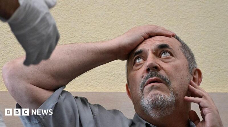 A man in a grey shirt holds his right arm to his forehead as he looks at a man out of the picture who is wearing a medical glove
