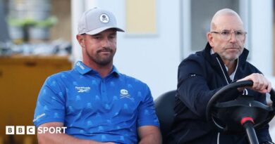 Bryson DeChambeau is driven on a buggy by an Open rules official