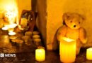 A white teddy bear sits against a cream wall with three candles in front of it. There are a collection of about a dozen tealights to the right of the teddy.