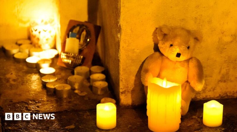 A white teddy bear sits against a cream wall with three candles in front of it. There are a collection of about a dozen tealights to the right of the teddy.