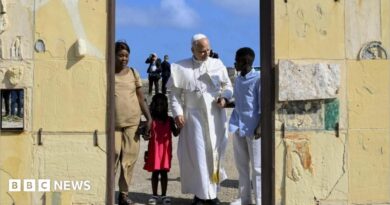 Pope Leo holds hands with a girl wearing a red dress and a man in blue shirt and white trousers. A women in a beige outfit stands alongside them at a doorway. Behind them are blue skies and men dressed in black