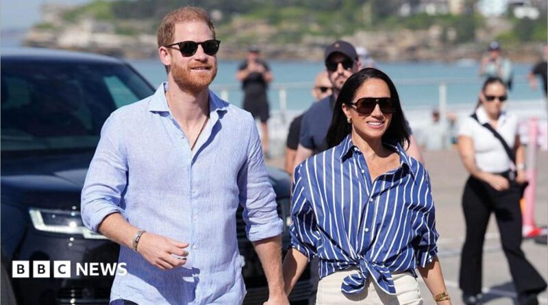 Prince Harry, Duke of Sussex, and his wife Meghan, the Duchess of Sussex walking hand in hand, wearing blue shirts and sunglasses.