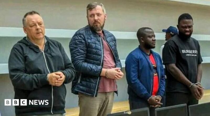 Four handcuffed men standing in front of a row of confiscated laptops. Lev Lakshtanov, Igor Ratchin, Amor Carlos Tomé and Francisco Oliveira (left to right)