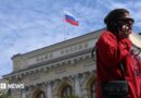 A woman wearing a red jumper and sunglasses, as well as a little bag on her right shoulder, talks on the phone in front of an exterior view of the Central Bank of the Russian Federation building on top of which the Russian flag can be seen
