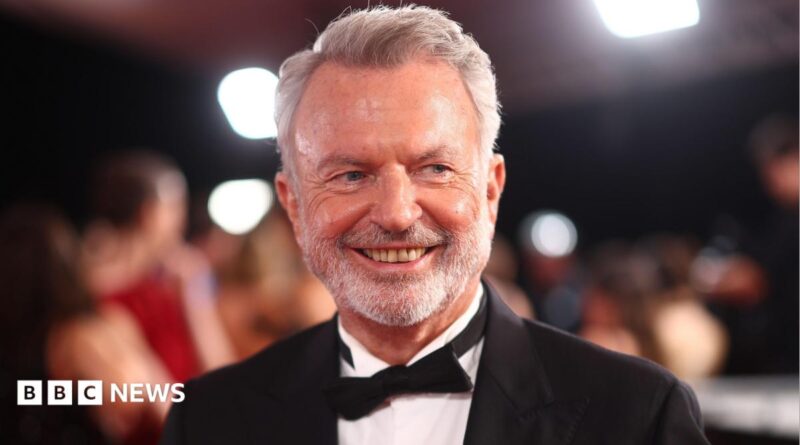 Graham Norton has white hair, a moustache and beard. He is wearing a dark-colour T-shirt and sits behind a microphone with a green muffler.