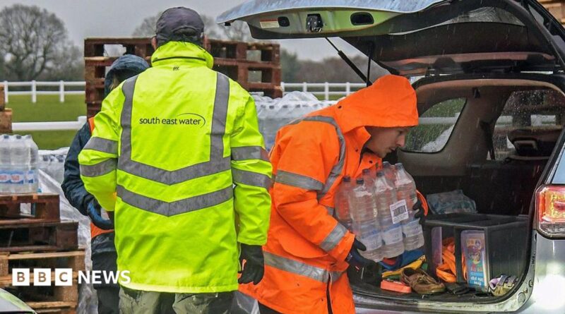 A man in a yellow hi-vis jacket with the word South East Water on the back. Another man, in an orange hi-vis jacket, is carrying a slab of water out of a car.
