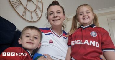 A woman in a white football top sits on a sofa smiling next to her children, wearing red England tops.