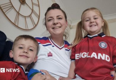 A woman in a white football top sits on a sofa smiling next to her children, wearing red England tops.