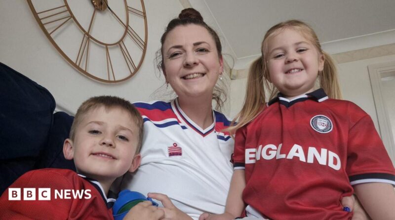 A woman in a white football top sits on a sofa smiling next to her children, wearing red England tops.
