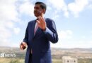 Khanna, who is dressed in a blue suit, white shirt and pink tie, raises his hand as he is photographed outdoors with a rural town landscape behind him