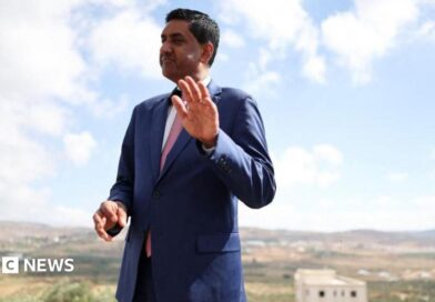 Khanna, who is dressed in a blue suit, white shirt and pink tie, raises his hand as he is photographed outdoors with a rural town landscape behind him