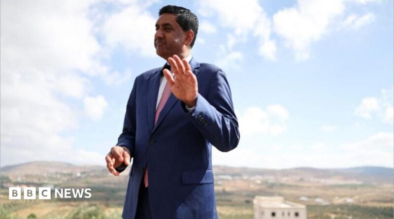 Khanna, who is dressed in a blue suit, white shirt and pink tie, raises his hand as he is photographed outdoors with a rural town landscape behind him