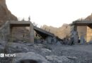 A damaged bridge with people standing on the remaining structure with mountains in the background, and rubble and rocks in the foreground.