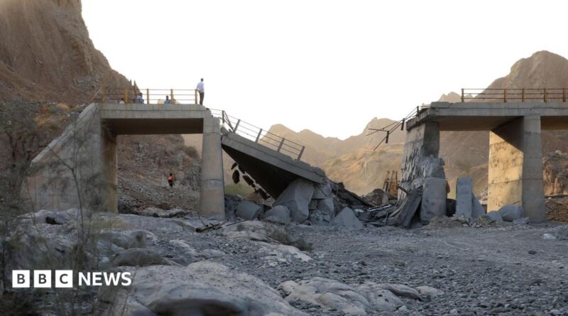 A damaged bridge with people standing on the remaining structure with mountains in the background, and rubble and rocks in the foreground.