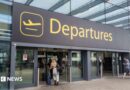 The departures entrance at Gatwick Airport with large yellow "Departures" signage above glass doors. Travellers with luggage can be seen entering the terminal building.