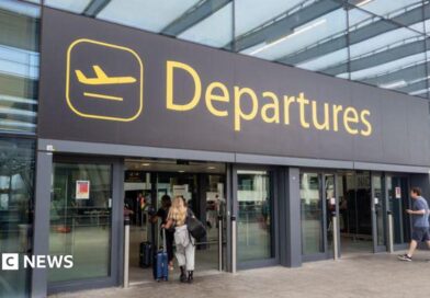 The departures entrance at Gatwick Airport with large yellow "Departures" signage above glass doors. Travellers with luggage can be seen entering the terminal building.