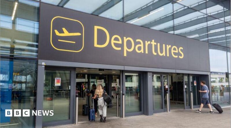 The departures entrance at Gatwick Airport with large yellow "Departures" signage above glass doors. Travellers with luggage can be seen entering the terminal building.