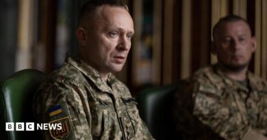 A man sits wearing a military uniform while another uniformed man looks on in the background