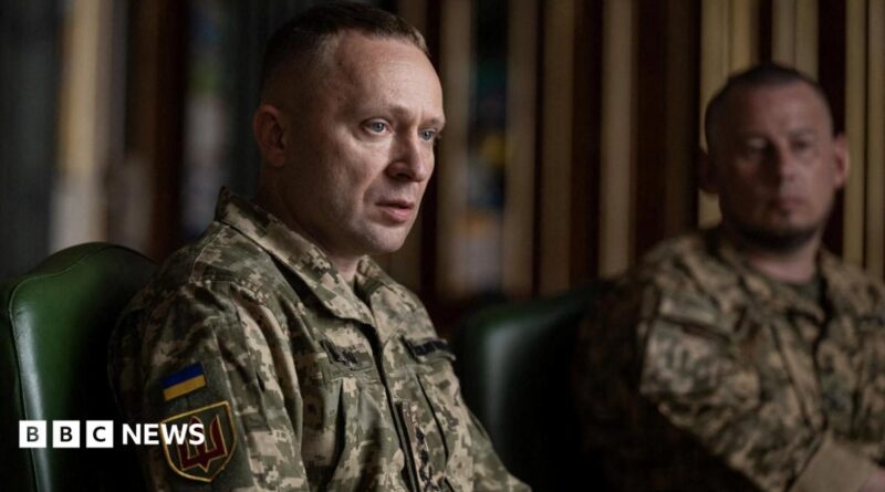 A man sits wearing a military uniform while another uniformed man looks on in the background