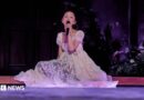 Ariana Grande sits on the stage, surrounded by flowers, as she sings on her Eternal Sunshine tour