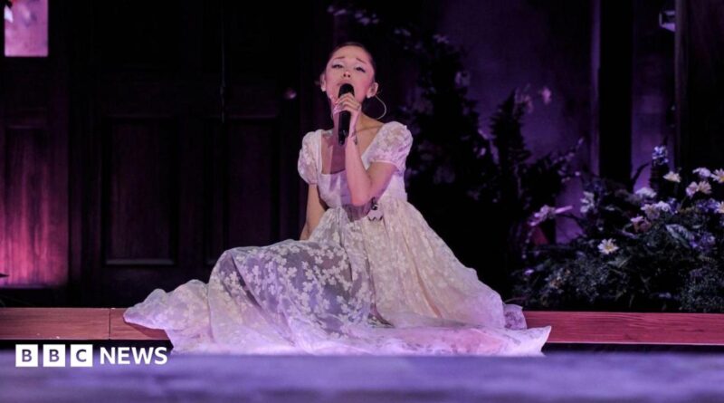 Ariana Grande sits on the stage, surrounded by flowers, as she sings on her Eternal Sunshine tour