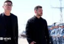 Andy Burnham and Volodymyr Zelensky walk next to each other at a naval base. Both men are dressed in black and have short, dark hair. Burnham is wearing glasses. Military personnel can be seen in the background.