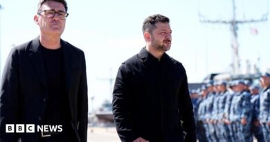 Andy Burnham and Volodymyr Zelensky walk next to each other at a naval base. Both men are dressed in black and have short, dark hair. Burnham is wearing glasses. Military personnel can be seen in the background.