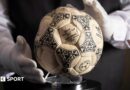A person wearing white gloves places the football used in the 1986 World Cup quarter-final on a stand