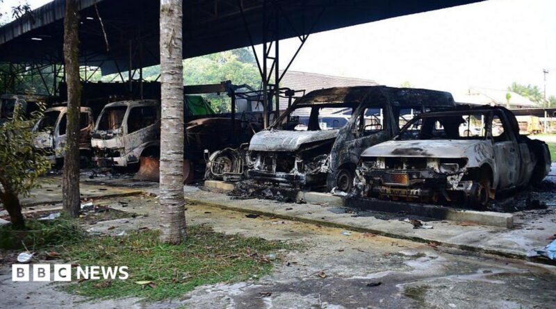 Five burned out vehicles are lined up under a tin roof structure