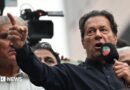 Imran Khan wearing a black button-up shirt and speaking into a microphone, points dramatically. Behind him are other men, and a television camera