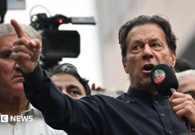 Imran Khan wearing a black button-up shirt and speaking into a microphone, points dramatically. Behind him are other men, and a television camera