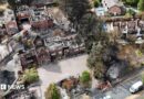 An aerial image shows several houses destroyed by fire, with their roofs gone and wreckage visible within. There is a line of dead trees next to the houses and areas of scorched earth. A fire engine is parked by one of the houses.