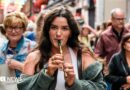 A woman with long brown hair, playing a tin whistle. There is a crowd of people behind her. 