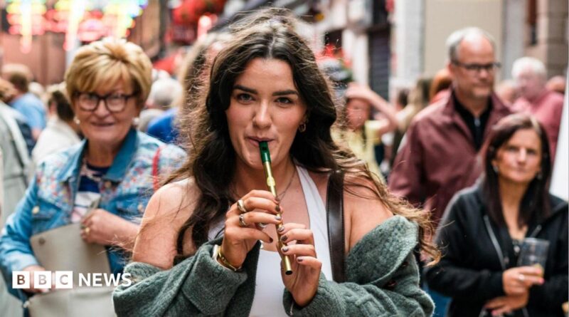 A woman with long brown hair, playing a tin whistle. There is a crowd of people behind her.